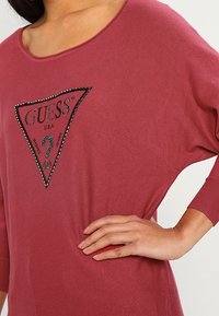 Camicia over-size bordeaux con scollatura ampia, caratterizzata da un logo triangolare contornato di nero con strass. Tessuto morbido e vestibilità rilassata.
