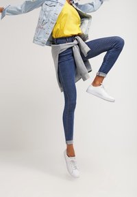 Donkerblauwe denim jeans met een slim fit, gecombineerd met een gele t-shirt en een lichtblauwe denim jacket. Witte sneakers en een grijze trui om de taille gebonden.