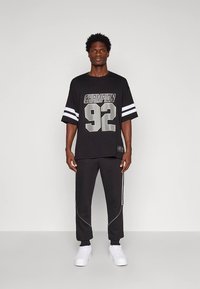 T-shirt preta de manga curta com gráfico "Champion 92" e riscas brancas nas mangas; combinada com calças de treino pretas com detalhes brancos. Ténis brancos.