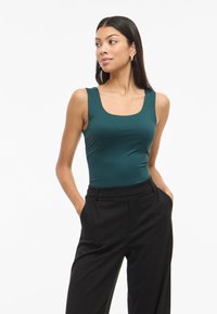 Body in tonalità turchese, aderente, con scollo quadrato e ampie spalline, abbinato a pantaloni neri su misura a vita alta.