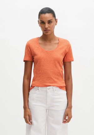 Femme aux cheveux foncés attachés, portant un t-shirt orange à manches courtes et un pantalon blanc, debout devant un fond blanc uni.