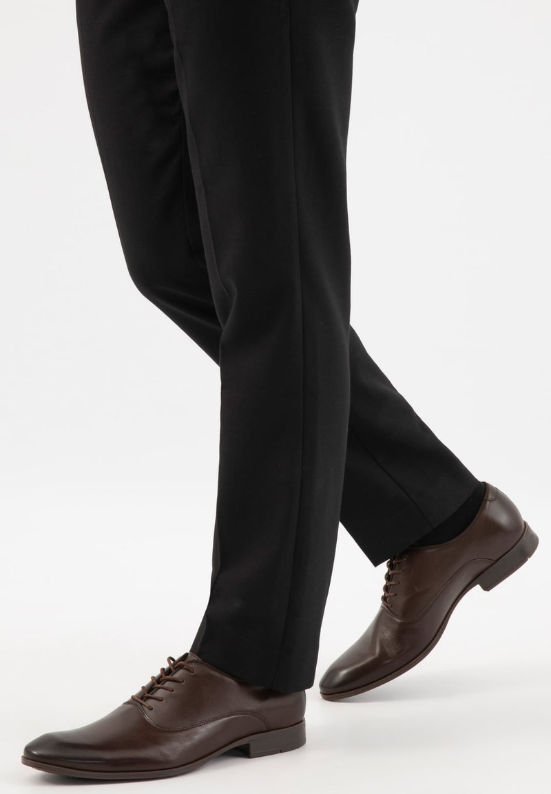 Jambes portant un pantalon de costume noir et des chaussures habillées en cuir marron foncé à lacets sur un fond blanc uni.