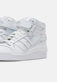 Zapatillas altas blancas de cuero sintético con una textura suave, que cuentan con una correa de Velcro, un diseño de cordones y un logotipo en relieve.