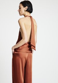 Satin-Jumpsuit in Rostbraun mit Neckholder-Ausschnitt, offenem Rücken, weitem Bein und Seitentaschen. Verfügt über einen fließenden Stoff und eine glatte Textur.