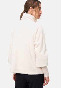 Veste en polaire blanche et duveteuse avec un col haut, des manches larges et des poignets côtelés. Texture lisse et apparence douce, idéale pour un style décontracté.