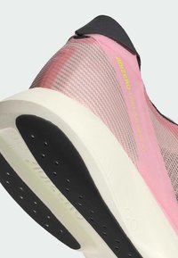 Primer plano del talón de una zapatilla de correr rosa y negra con suela blanca, parte superior de malla transpirable y texto amarillo "ADIZERO TAKUMI SEN" en el lateral.