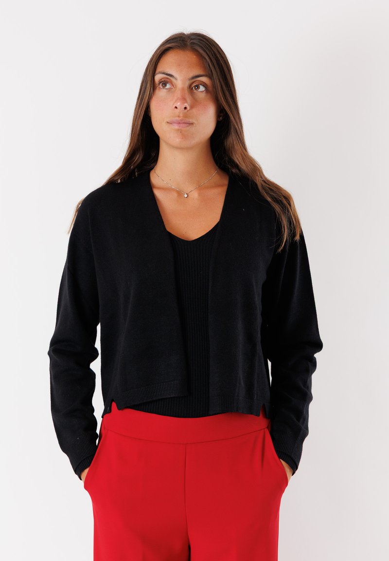 Cardigan nero corto realizzato in materiale morbido, con maniche lunghe e design aperto, abbinato a un top nero a costine e pantaloni rossi.