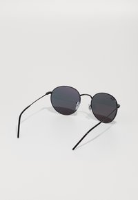 Ochelari de soare rotunzi cu lentile închise la culoare și un cadru subțire din metal negru. Au pernuțe pentru nas transparente și brațe subțiri negre.