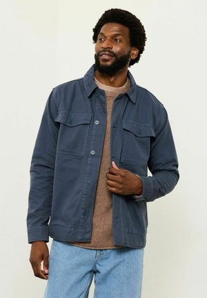 Marineblå lærredjakke med knaplukning, to brystlommer og krave, båret over en beige striktrøje og lyseblå jeans.