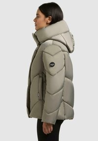 Leichte graue Steppjacke mit hohem Kragen und Kapuze. Sie verfügt über eine Chevron-Steppung, zwei seitliche Taschen und ein Logo-Patch am Ärmel.