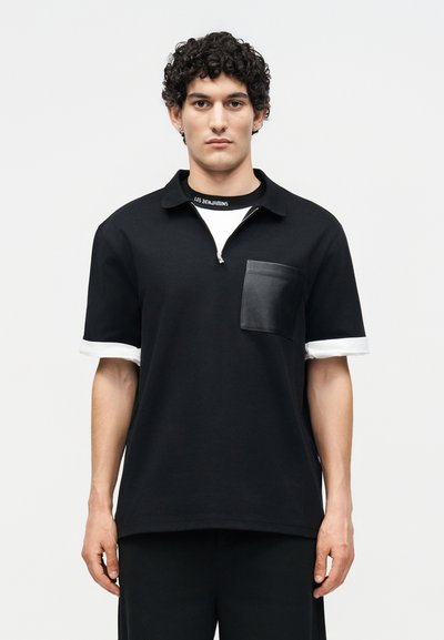 Νεαρός άνδρας με σγουρά μαλλιά, που φοράει μαύρο κοντομάνικο πουκάμισο με λευκές μανσέτες και δερμάτινη τσέπη στο στήθος, κοιτάζοντας μπροστά με απλό φόντο.