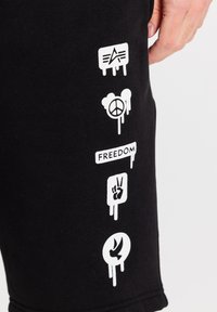 Sorte sweatpants med hvide trykte designs, herunder symboler for fred, frihed og en fugl, med en glat bomuldsstruktur.