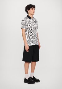 Pólo de mangas curtas com um padrão de texto em preto e branco, gola preta, combinado com calções soltos pretos e sapatos pretos de deslizar.