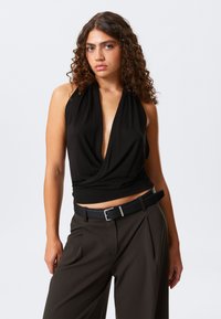 Top senza maniche nero con scollatura a V profonda e parte anteriore drappeggiata, abbinato a pantaloni marroni scuri a vita alta e una cintura nera.