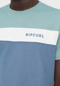Κοντομάνικο T-shirt σε τυρκουάζ, λευκό και μπλε. Διαθέτει οριζόντια μπλοκ χρωμάτων και το κείμενο «RIPCURL» σε ανοιχτό μπλε χρώμα στο στήθος. Κατασκευασμένο από βαμβάκι.