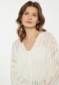 Fehér, kötött kardigán gyémántmintával, V-nyakkivágással és gombos záródással. Hosszú ujjakkal rendelkezik, nyitott szövésű, így légáteresztő.