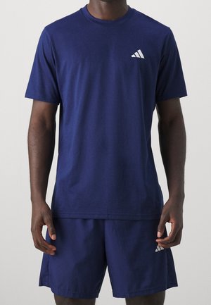 Mężczyzna w granatowej sportowej koszulce Adidas i pasujących szortach stoi na jednolitym tle.