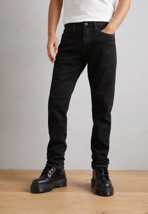 Jeans de mezclilla negra con un ajuste slim, que presentan costuras en contraste y bolsillos laterales, combinados con botas de combate negras gruesas sobre un suelo de madera.