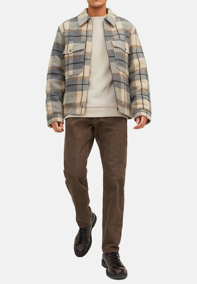 Geruite wollen jas in grijs, beige en blauwe tinten met twee borstzakken; gedragen over een lichte trui en gecombineerd met bruine corduroy broeken en zwarte laarzen.