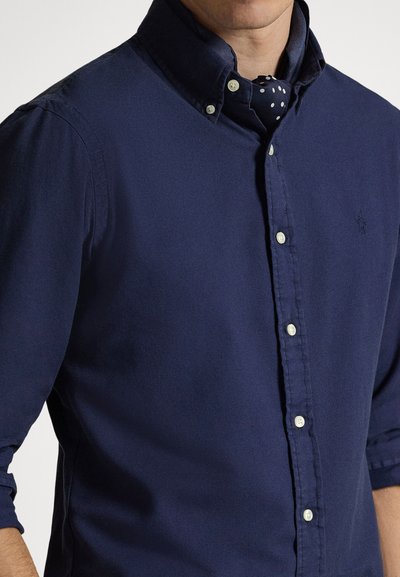Κοντομάνικο πουκάμισο σε σκούρο μπλε χρώμα, από ελαφρύ ύφασμα με γιακά, διακοσμημένο με λευκά κουμπιά και διακριτική υφή. Συνδυάζεται με γραβάτα που έχει πουά σχέδιο.