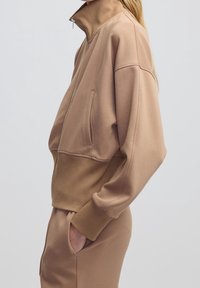 Persona con una giacca beige con zip, polsini e vita a costine, con una mano nella tasca dei pantaloni beige abbinati, vista di lato.