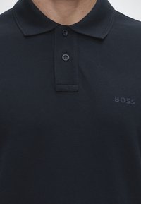 Mustast poloülikonnast lähivaade, millel on kaks nööpi ja vasakul rinnal tikitud "BOSS" logo.