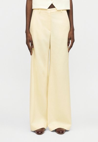 Personne portant un pantalon crème taille haute à jambes larges et un gilet court assorti, debout les bras détendus le long du corps.
