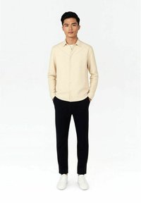 Ljusbeige skjorta med knappar, mjukt tyg. Svarta byxor, slim fit. Vita sneakers. Modellen står mot en neutral bakgrund med händerna i fickorna.
