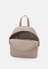 Světle taupe batoh s hladkou texturou, hlavní přihrádkou na zip, zlatým kováním a vnitřní kapsou. Má dvojité horní úchyty.