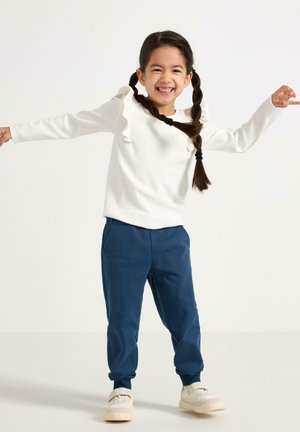 Valkoinen pitkähihainen paita, jossa röyhelökoristeita, yhdistetty sinisiin kapeneviin housuihin. Mallilla on valkoiset tennarit ja hänen hiuksensa ovat kahdella letillä.