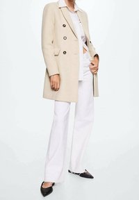 Cappotto doppiopetto beige con bottoni neri, indossato sopra una camicia bianca, abbinato a pantaloni bianchi a vita alta e décolleté nere con punta appuntita.