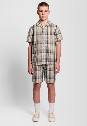 Tienerjongen staat met het gezicht naar voren, draagt een beige en marineblauw geruit kortärmelig overhemd met knopen en bijpassende korte broek, witte sokken en crèmekleurige sneakers.