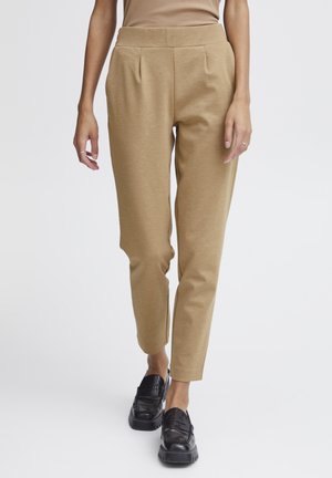 Beige, schmal zulaufende Hose mit glatter Textur, die vorne Falten und einen hohen Taillenbund hat, kombiniert mit schwarzen Mokassins.