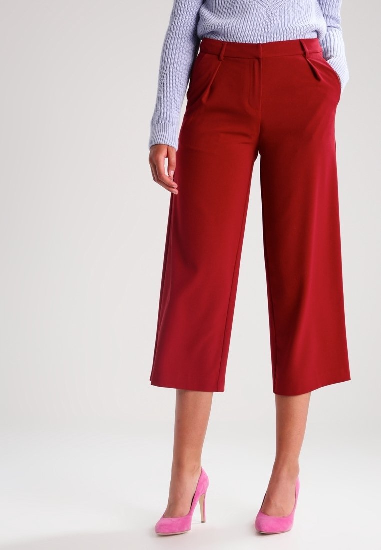 Femme portant un pantalon rouge large raccourci, un pull violet clair et des chaussures à talons hauts roses, debout les mains dans les poches.