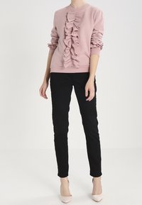Sudadera rosa con un frente con volantes, cuello redondo y mangas fruncidas, combinada con pantalones negros ajustados y zapatos de color claro.