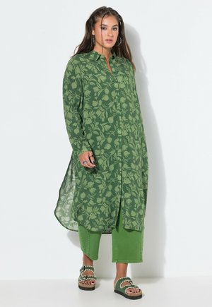 Kvinde iført en lang grøn blomsterknap-op-skjorte, grønne korte bukser og grønne sandaler med åbne tæer, stående mod en hvid baggrund.
