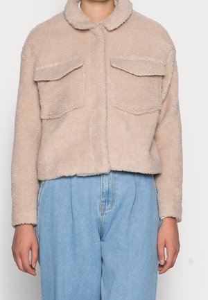 Chaqueta de piel de oveja sintética beige con cuello, dos bolsillos frontales y un diseño corto. Llevada sobre pantalones de mezclilla de tiro alto en azul claro.