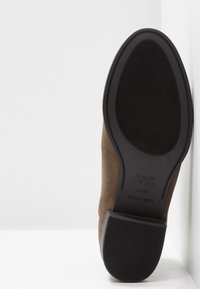 Une chaussure en daim marron posée sur le côté montrant la semelle en caoutchouc noir avec la texture du talon et de la semelle ainsi que les détails de la taille.