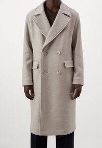 Manteau double boutonnage en mélange de laine beige avec larges revers, trois boutons à l'avant et deux poches latérales, présentant une silhouette droite.