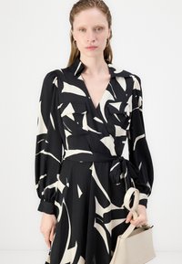 Black dress with white abstract patterns, long sleeves, collar, and tie waist. Features two front pockets. Model holds a beige bag.