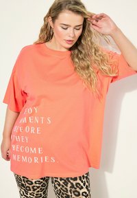 Camiseta de gran tamaño en color coral con texto blanco que dice "Disfruta los momentos antes de que se conviertan en recuerdos." Combinada con leggings de estampado de leopardo.