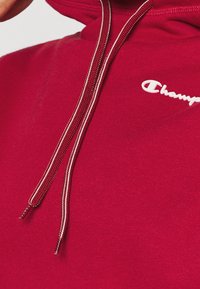 Czerwona bluza z kapturem, wykonana z miękkiego materiału, z regulowanym kapturem. Posiada kontrastowe, białe logo oraz akcenty w postaci sznurków. Gładka faktura i wykończenie.