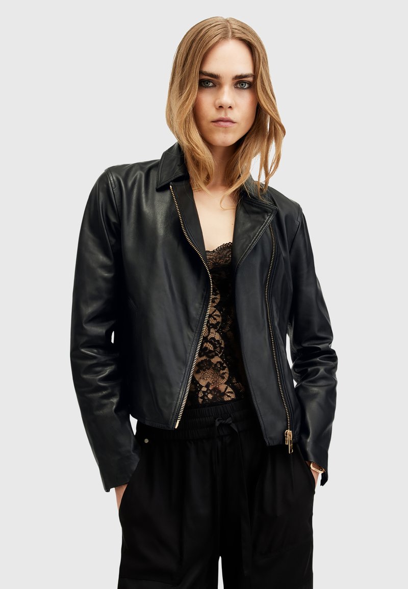 Giacca di pelle nera con silhouette aderente, zip frontale e colletto a punte. Abbinata a un top in pizzo e pantaloni neri.