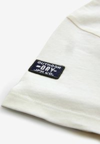 Kortærmet hvid bomulds skjorte med et blåt rektangulært broderet mærke med teksten "OUTDOOR DRY JPN CO." på ærmet.