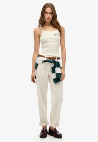Witte geribde tanktop met kantafwerking, groen bedrukt logo, gecombineerd met losse beige broek en een groen gestreepte tailleband. Donkere instapschoenen.