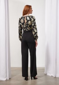 Mono negro con cintura ajustada, mangas largas con estampado floral en dorado y negro, y un detalle de espalda con abertura en forma de llave. Textura suave y sedosa.