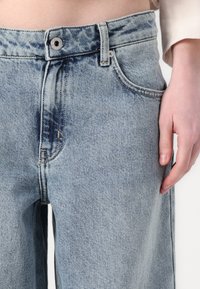 Svetlo modri kavbojke z visokim pasom, sprednjim gumbom in vidnimi stranskimi žepi. Tkanina ima rahlo teksturiran zaključek.