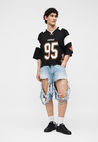 Jeune homme portant un maillot de sport noir et blanc numéro 95 et un short en jean bleu clair très abîmé avec de grandes déchirures.