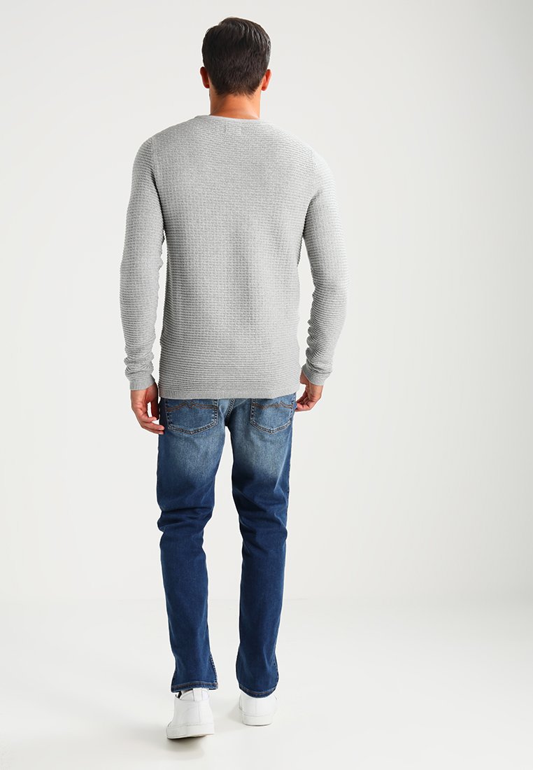 Grauer gestrickter Pullover mit langen Ärmeln, kombiniert mit blauen Jeans. Das Model steht mit dem Rücken zu einem hellen Hintergrund und trägt weiße Sneaker.