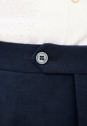 Nahaufnahme eines Bunds navyblauer Hose mit einem einzelnen runden irisierenden Knopf, kombiniert mit einem strukturierten weißen Strickoberteil.